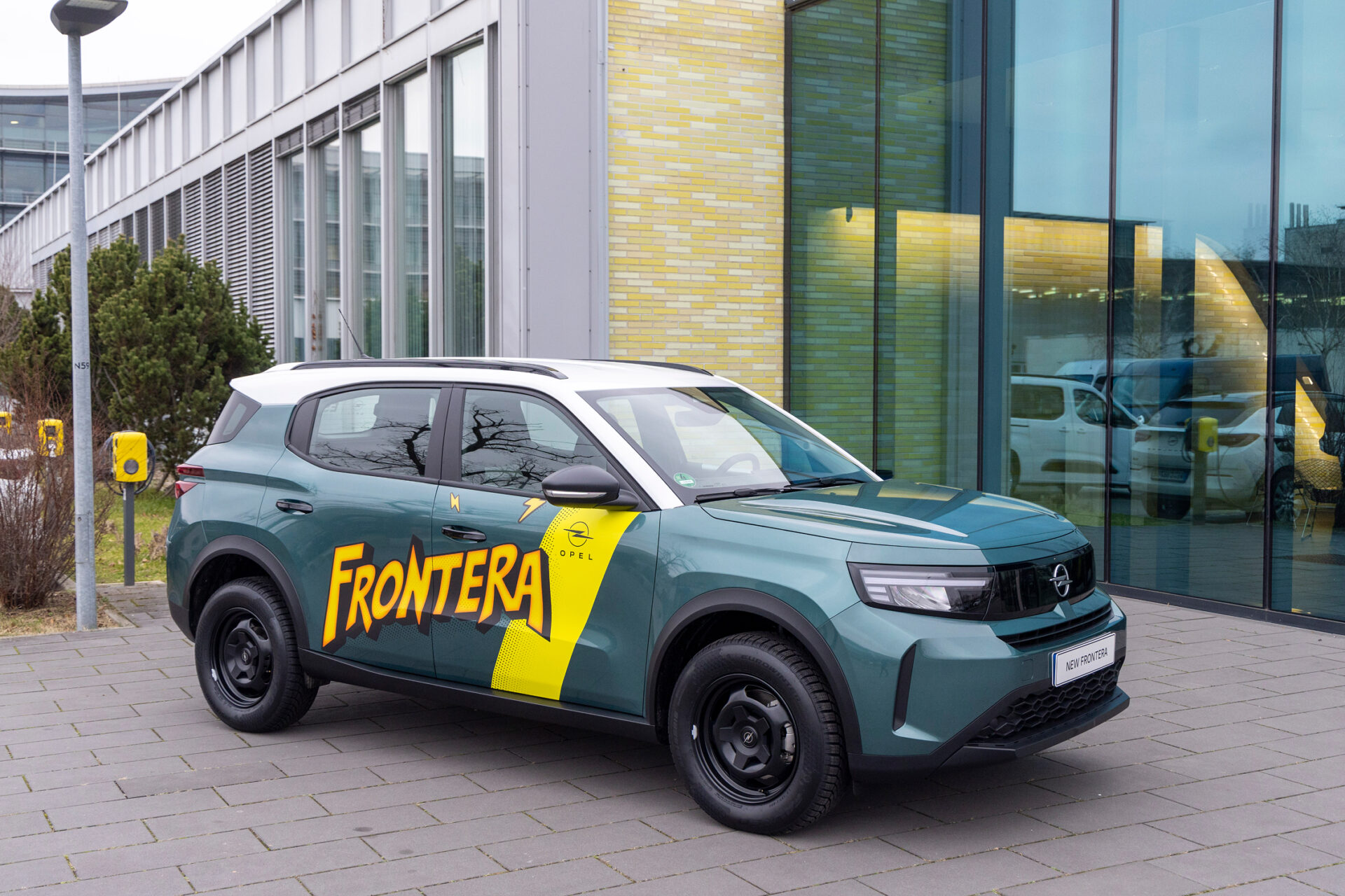 Grüner Opel Frontera mit gelber Comic-Beschriftung vor modernem Firmengebäude, klarer Fokus auf Fahrzeugdesign und Branding.