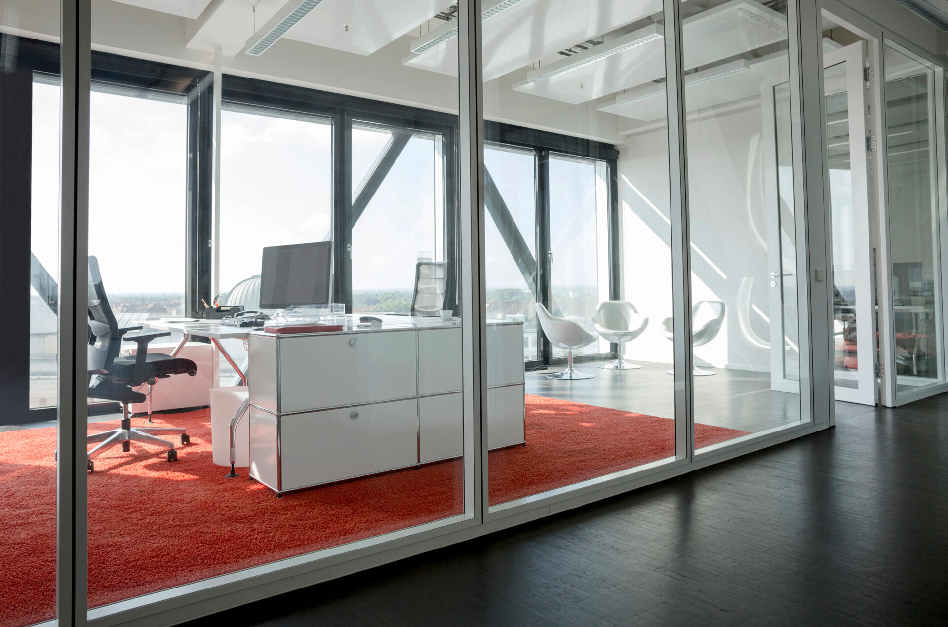 Modernes Büro mit Glaswänden, rotem Teppich, Schreibtisch, Bürostuhl und weißem Stauraum, im Hintergrund Besprechungsecke mit vier Sesseln.