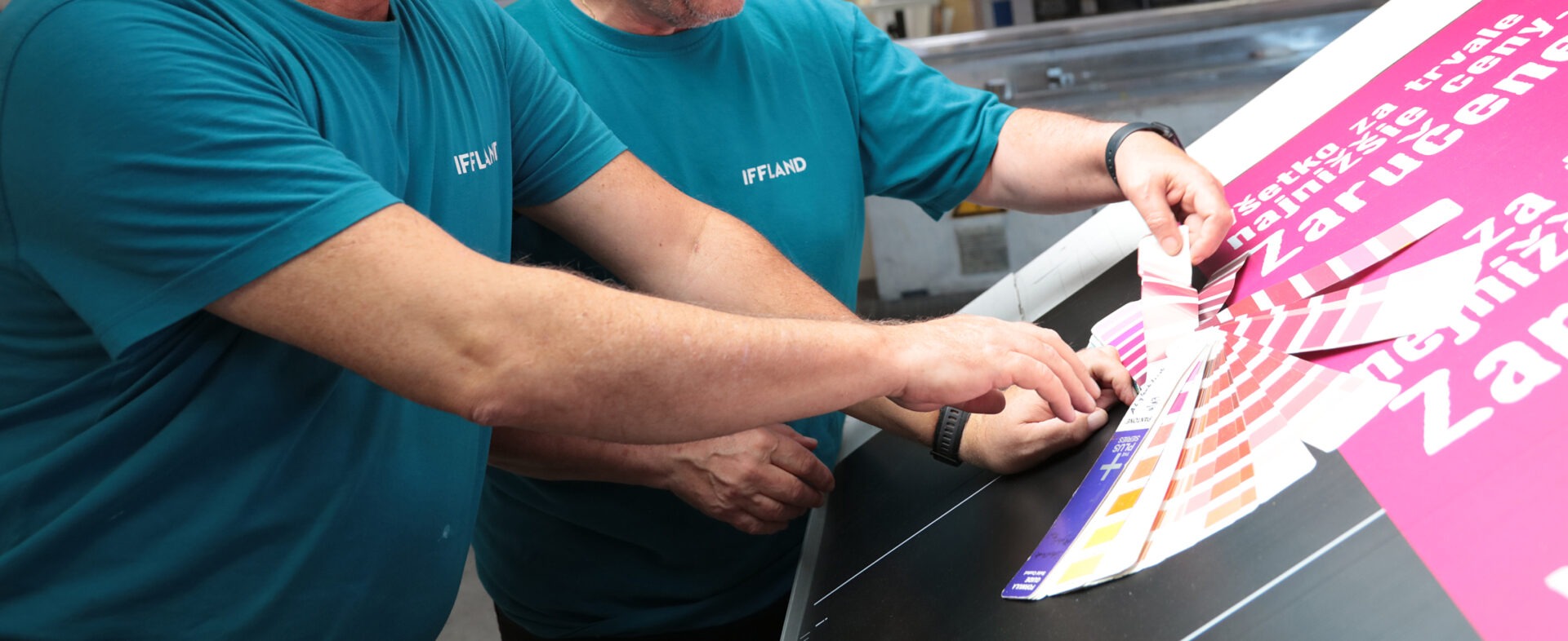 Zwei IFFLAND Mitarbeiter kontrollieren mit einem Pantone Farbfächer das fertige Druckprodukt zur Sicherstellung der Farbqualität.