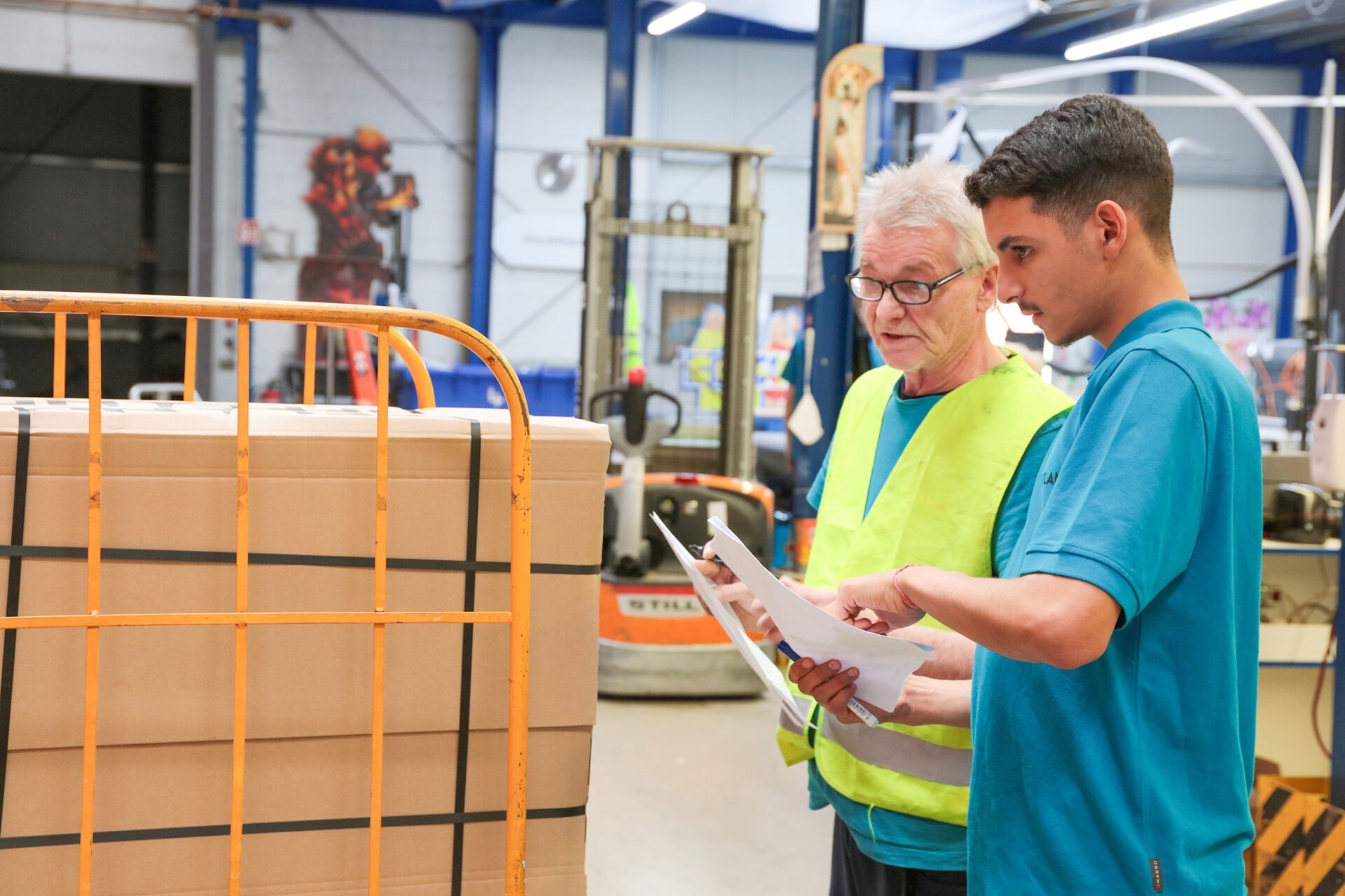 Junger Mitarbeiter bespricht sich im Lager mit Logistik-Experten. Beide haben Papierausdrucke in der Hand und begutachten ein großes zusammengeschnürtes Warenpaket.